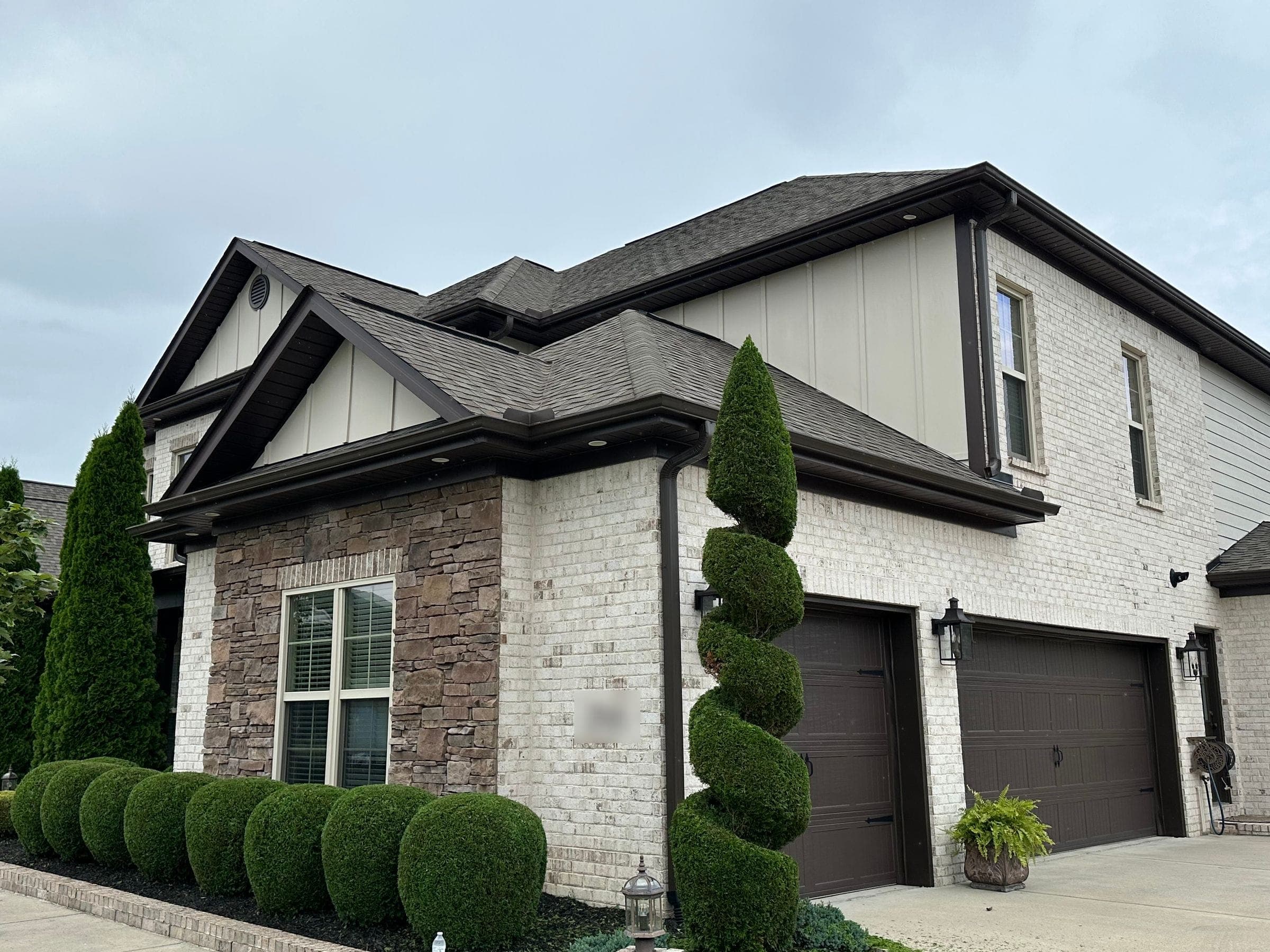 A large brick and stone home with a tall spiral topiary and a long row of round clipped boxwoods, trimmed that day with the clippings still on the drive.