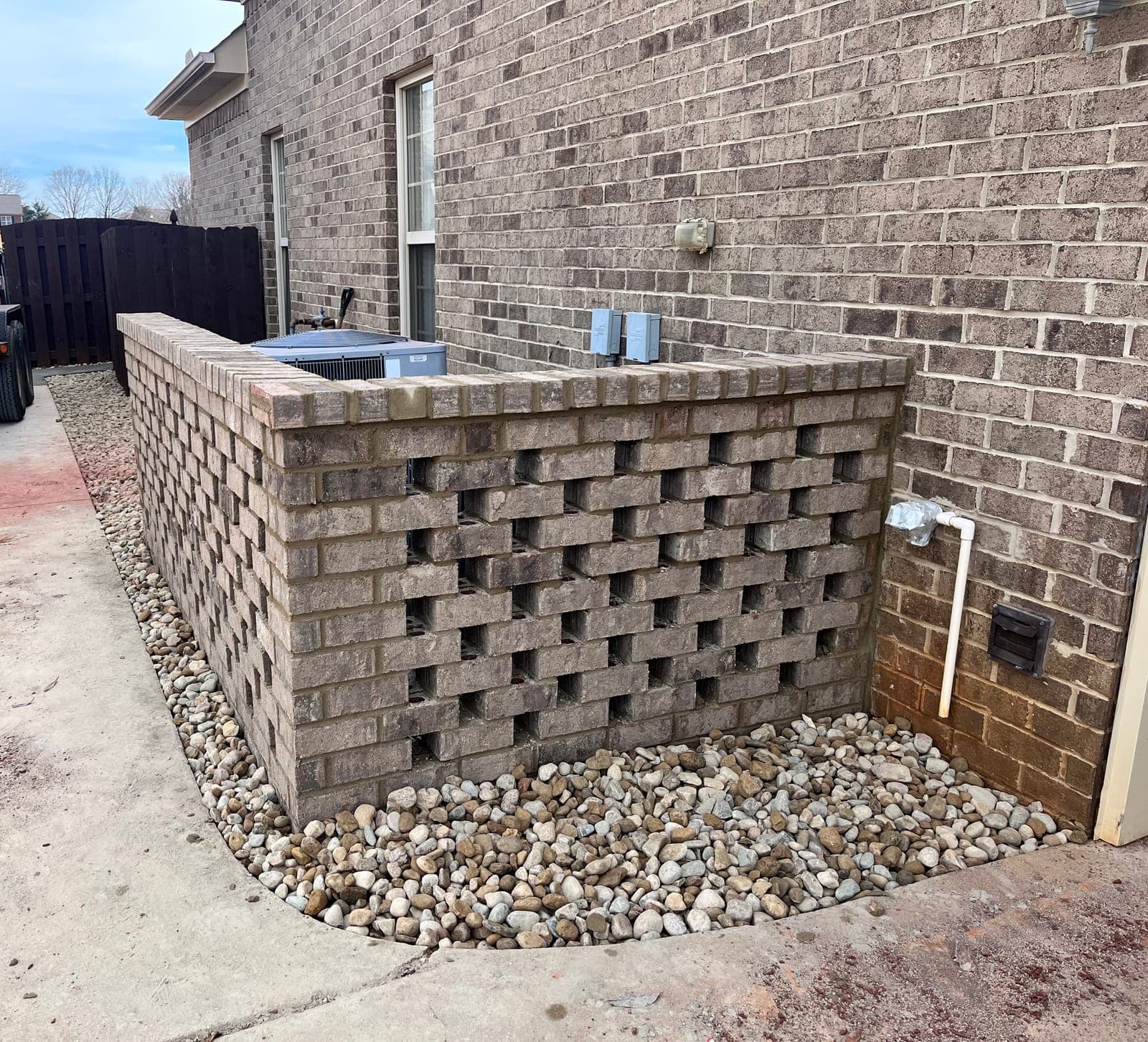 A patterned block screen wall built to hide an air conditioning unit, finished with a bed of river rock along its base.