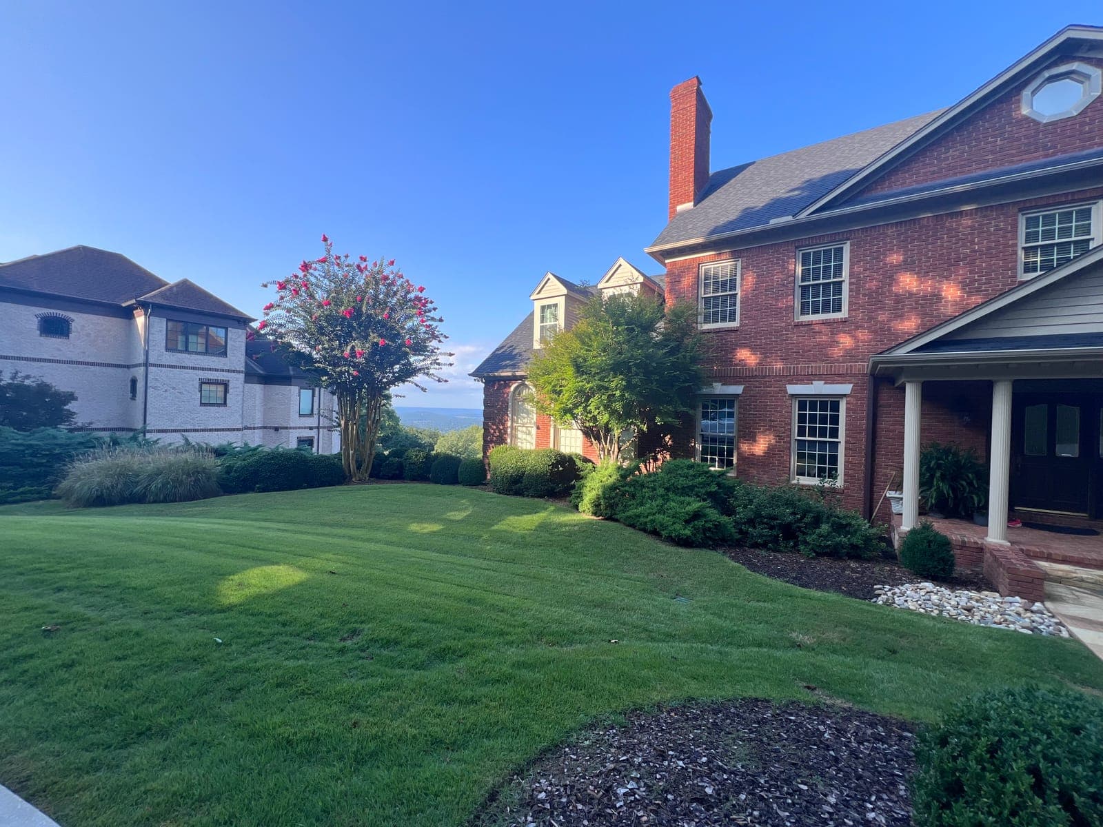 A freshly striped lawn wrapping the corner of a brick home, with a crepe myrtle in full bloom and a valley view beyond.