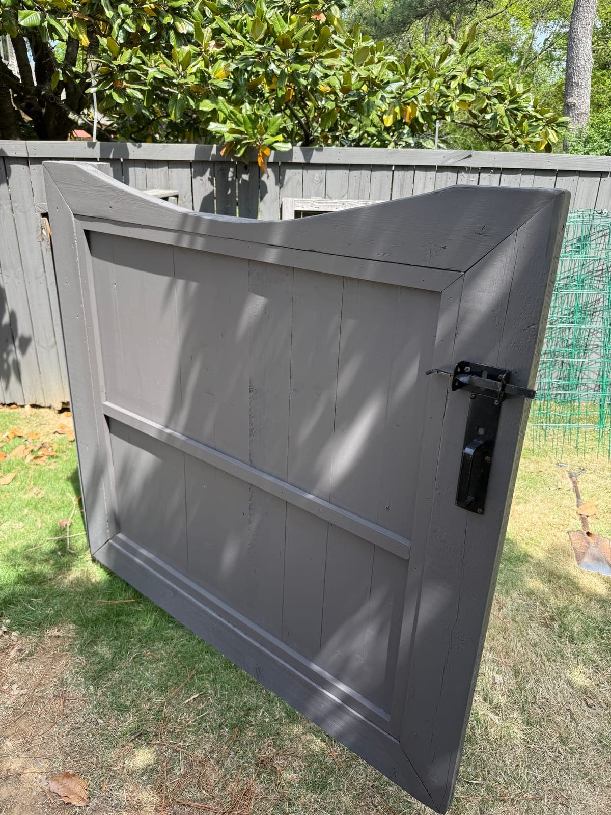 A rebuilt wood gate painted gray from its flat face, with a black thumb latch set at hand height under a magnolia.