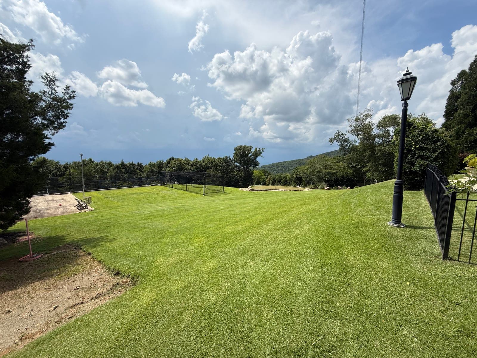 A freshly mown hillside lawn running out to a black iron fence and a lamp post, with a wooded ridge on the horizon.