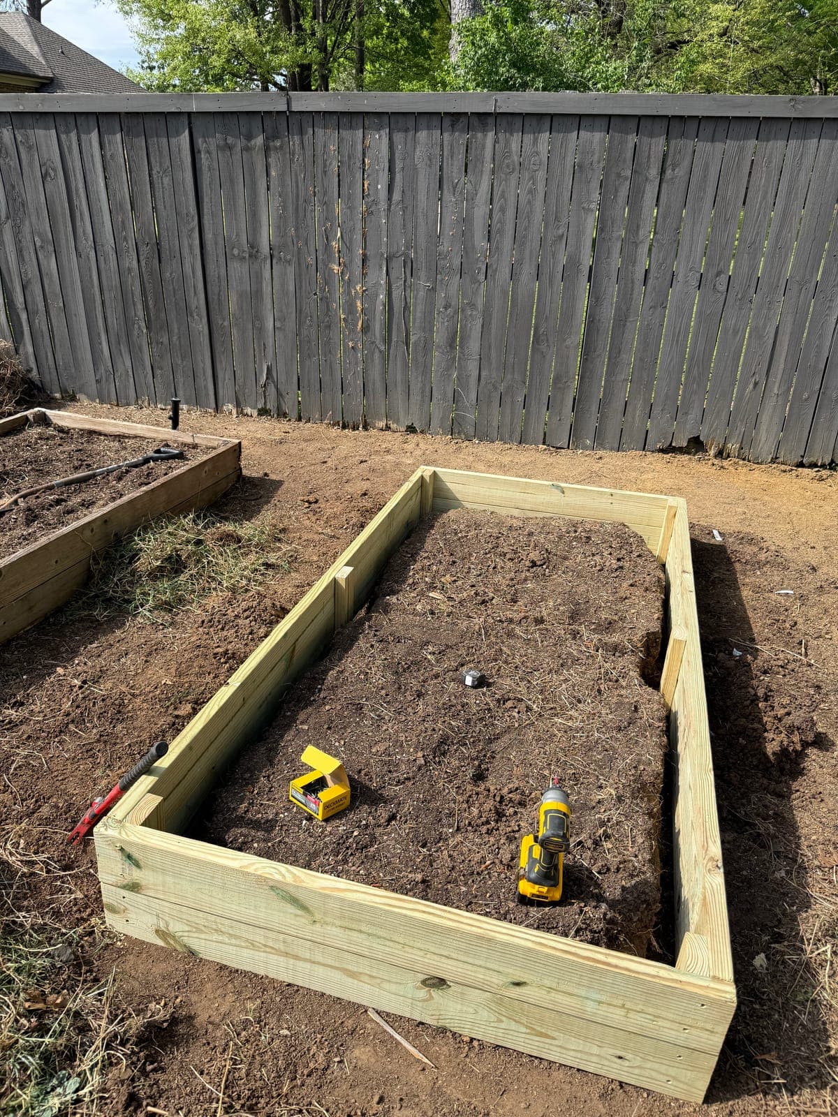 A new raised garden bed frame of pressure treated lumber built two courses high on turned soil, beside an older weathered bed.