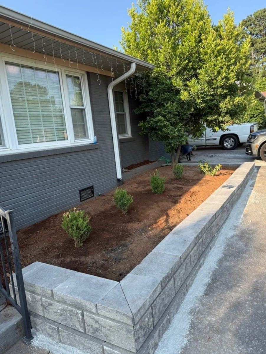 A new block retaining wall along a driveway, holding a freshly planted bed of young shrubs.