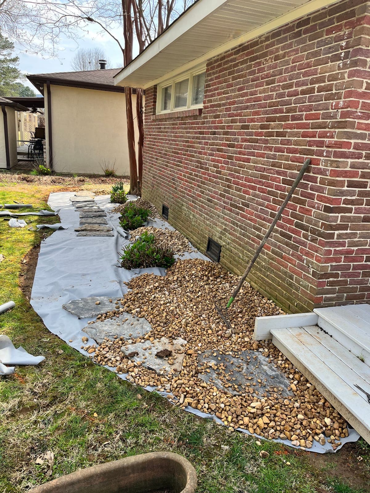 A river rock drainage bed laid over ground cloth along a brick foundation, planted with small shrubs between the stones.