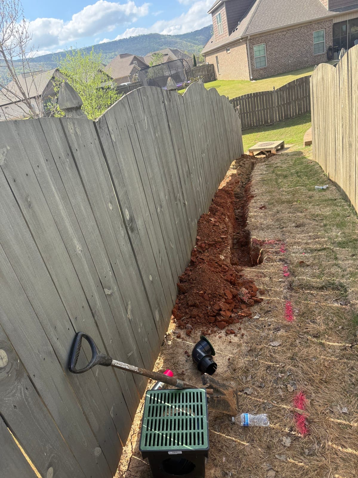 An open trench cut through red clay along a wood privacy fence, with a green catch basin and black pipe fittings staged on the grass beside it.