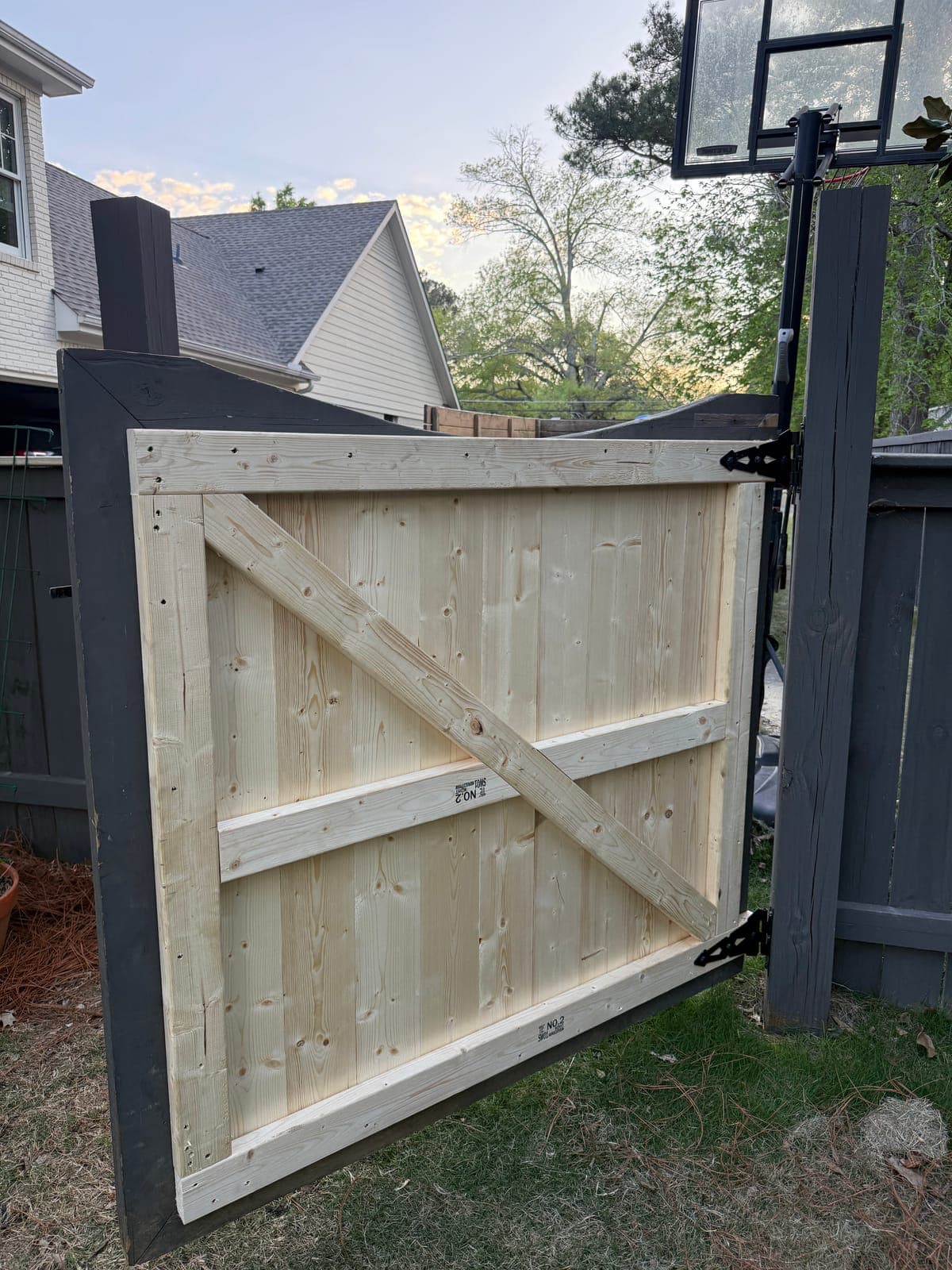 A newly built wooden gate of bare pine boards with a diagonal brace, hung on black strap hinges between dark stained fence posts.