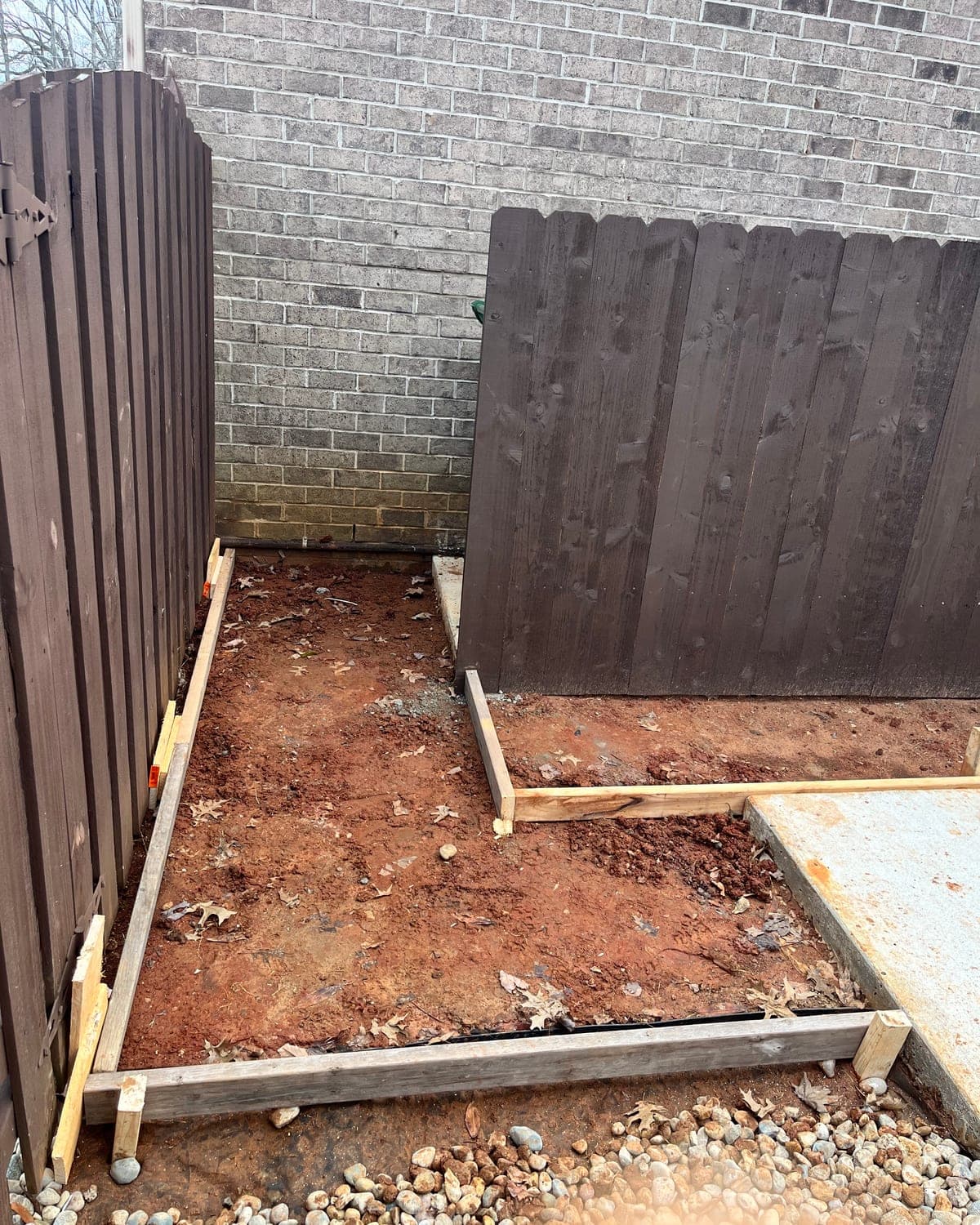 A narrow side yard of bare red clay between a brown fence and a brick wall, boxed in by fresh wood concrete forms.