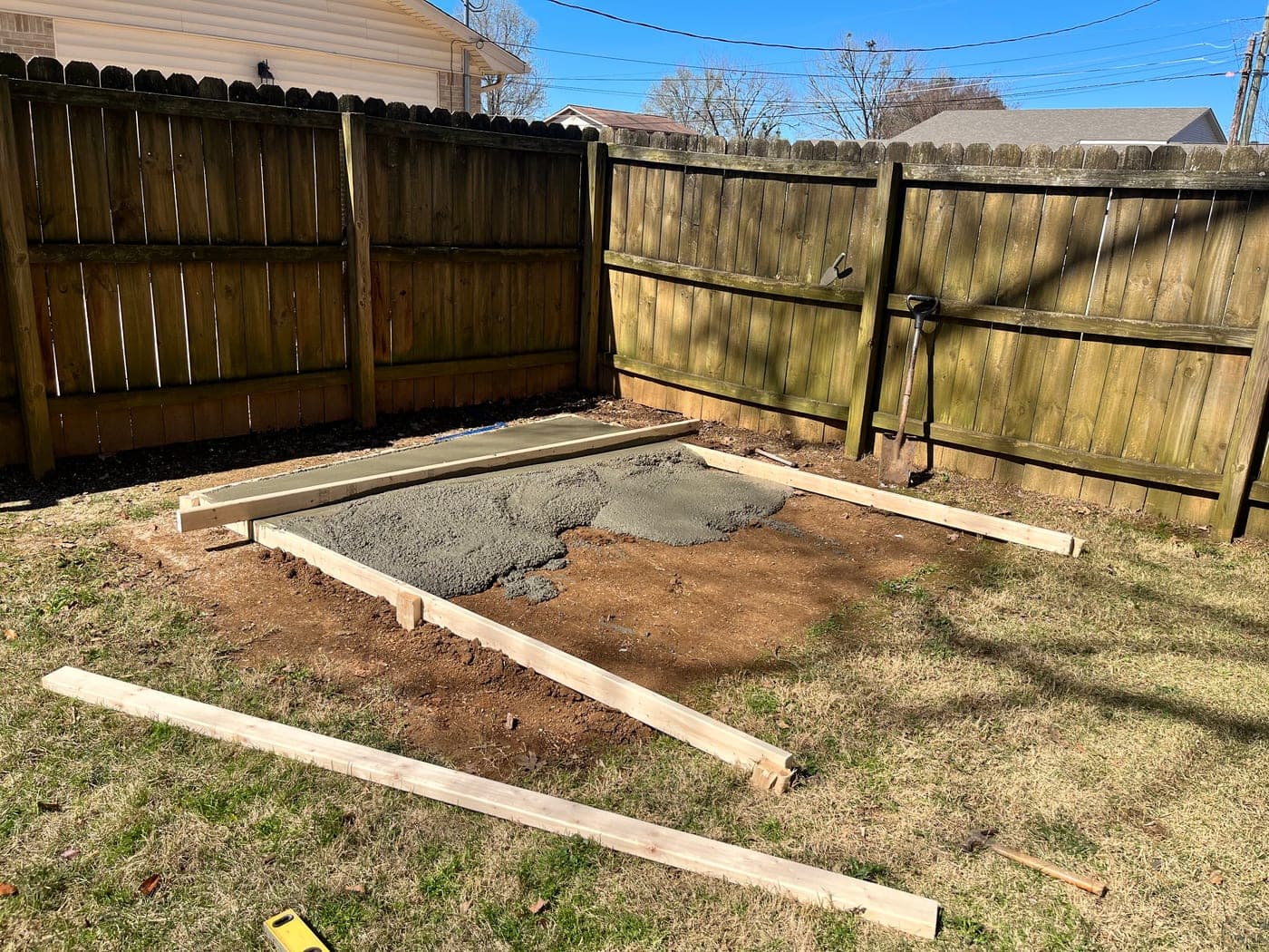 A back yard corner with the ground dug out and wood forms set, the first concrete going in along one edge.